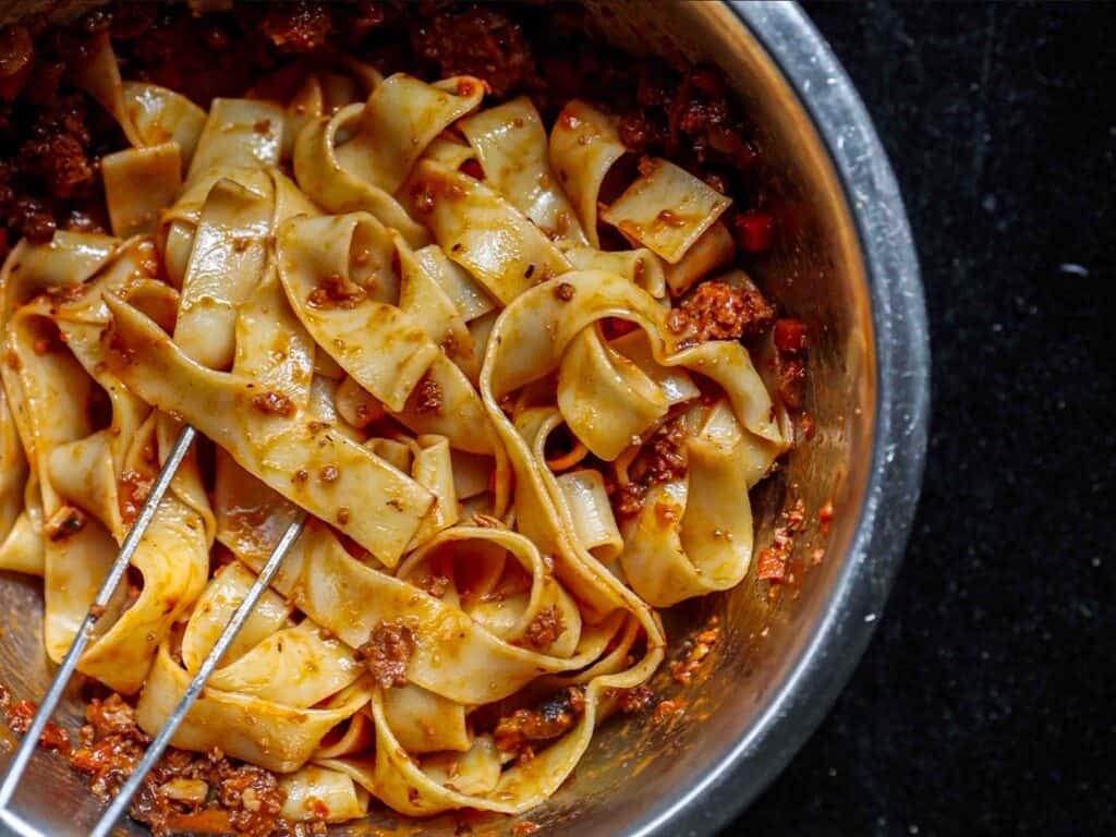 A bowl of wide, flat pasta noodles coated in a rich, red vegan bolognese sauce, with a pair of metal tongs resting in the dish, all set against a dark background.